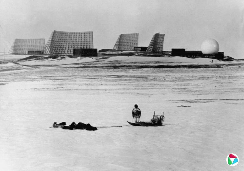 1966年,格陵兰岛北部,一名当地人带着他的狗拉雪橇,凝望着美国人建造的图勒空军基地。