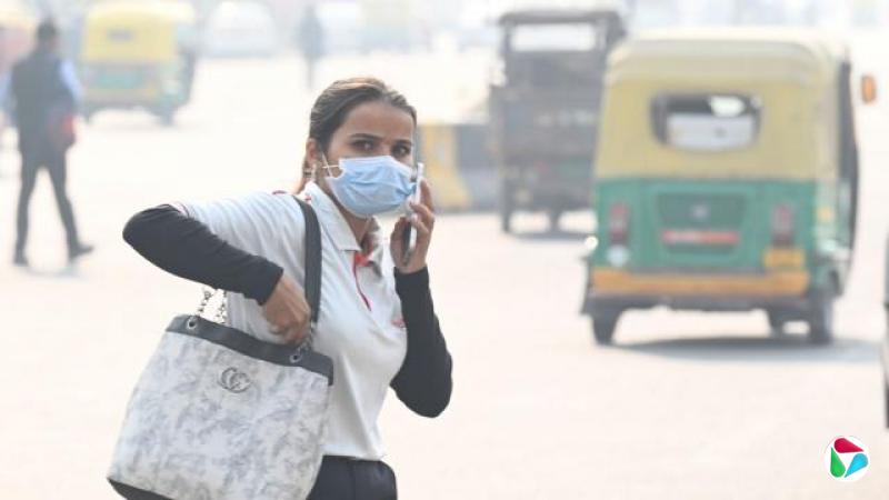 一名戴着口罩、在诺伊达(Noida)街头边走边打电话的女性,背景可见行驶中的三轮嘟嘟车。摄于2025年11月。