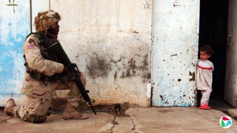 一名美国士兵在巡逻时守卫街道,他跪在水泥地上,枪口朝下。他穿着迷彩服。一个年轻女童站在建筑物角落看着他。