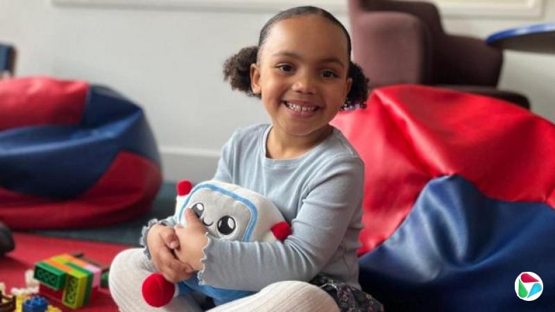 一名年幼的孩子正抱着AI玩具Gabbo。孩子坐在柔软座位与其他玩具之间,面向镜头