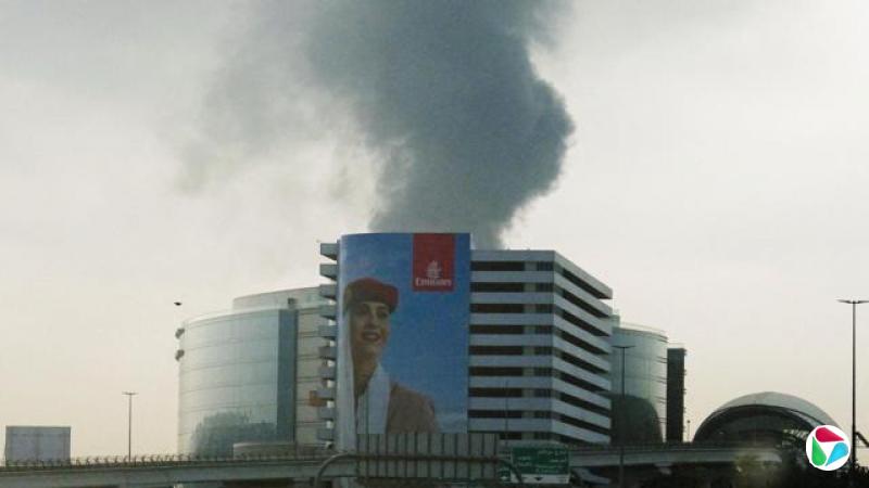 无人机袭击击中杜拜国际机场附近的一个油罐后,浓烟从汽车挡风玻璃升起。画面前景中,一张阿联酋航空的海报几乎覆盖了一栋建筑物的整个外墙,海报上是一位女性空服员。