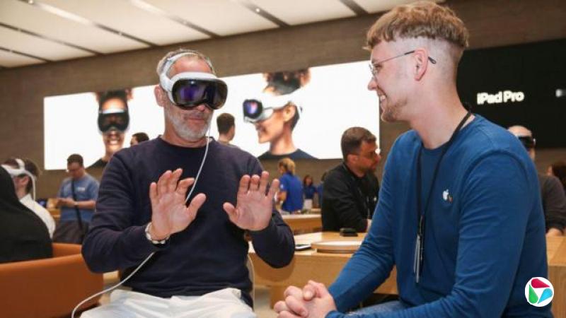 德国柏林某苹果专卖店内,一名男子坐着,头上戴着白色 Apple Vision Pro 耳机,耳机透过白色线连接。他双手伸向前方。一位苹果员工坐在他旁边,带着微笑。(2024年7月)