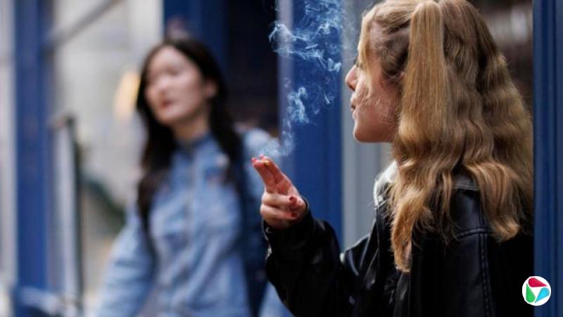 一位年轻女子在商店外抽烟。她留着金色长发,穿着一件皮夹克。
