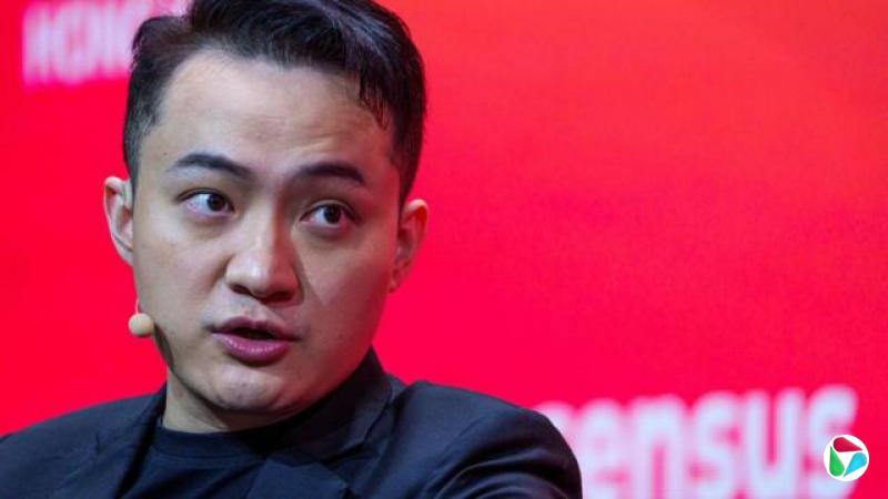 孙宇晨身穿黑色西装外套和黑色T恤,手持米色小麦克风和耳麦,站在红色背景前。