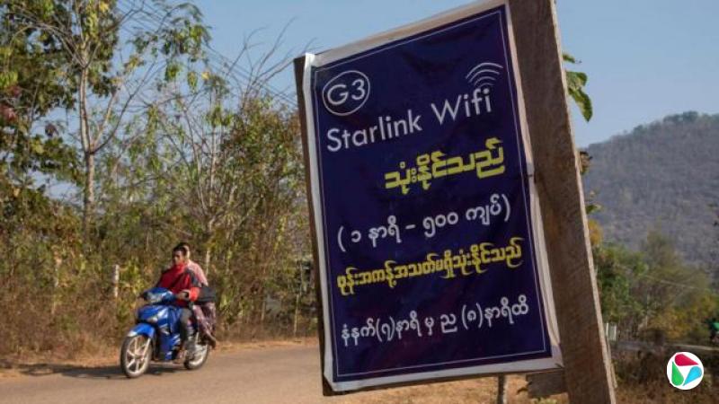 在缅甸克耶邦,一块贴在木板上的纸质告示以英文写着“Starlink Wifi”,并附有缅文说明。背景中可见两人骑着一辆蓝色电单车行驶在道路上,远处则是绿树覆盖的山丘。(16/2/2025)