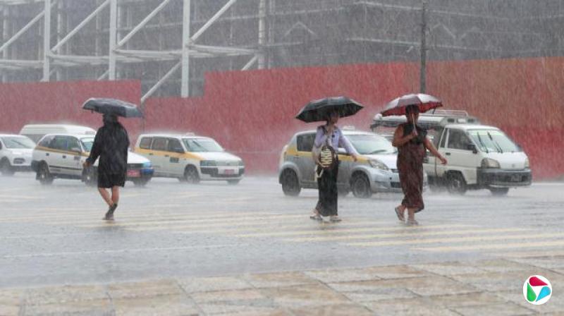 在缅甸,一条道路上有三人在大雨中行走,背景可见车辆,他们都撑着雨伞,雨水在行人过路处的路面上弹起