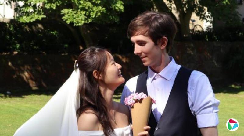 一对年轻夫妇身穿婚礼服饰在户外。两人互相凝视并微笑。新娘戴着头纱,手持一个装有鲜花的纸锥。